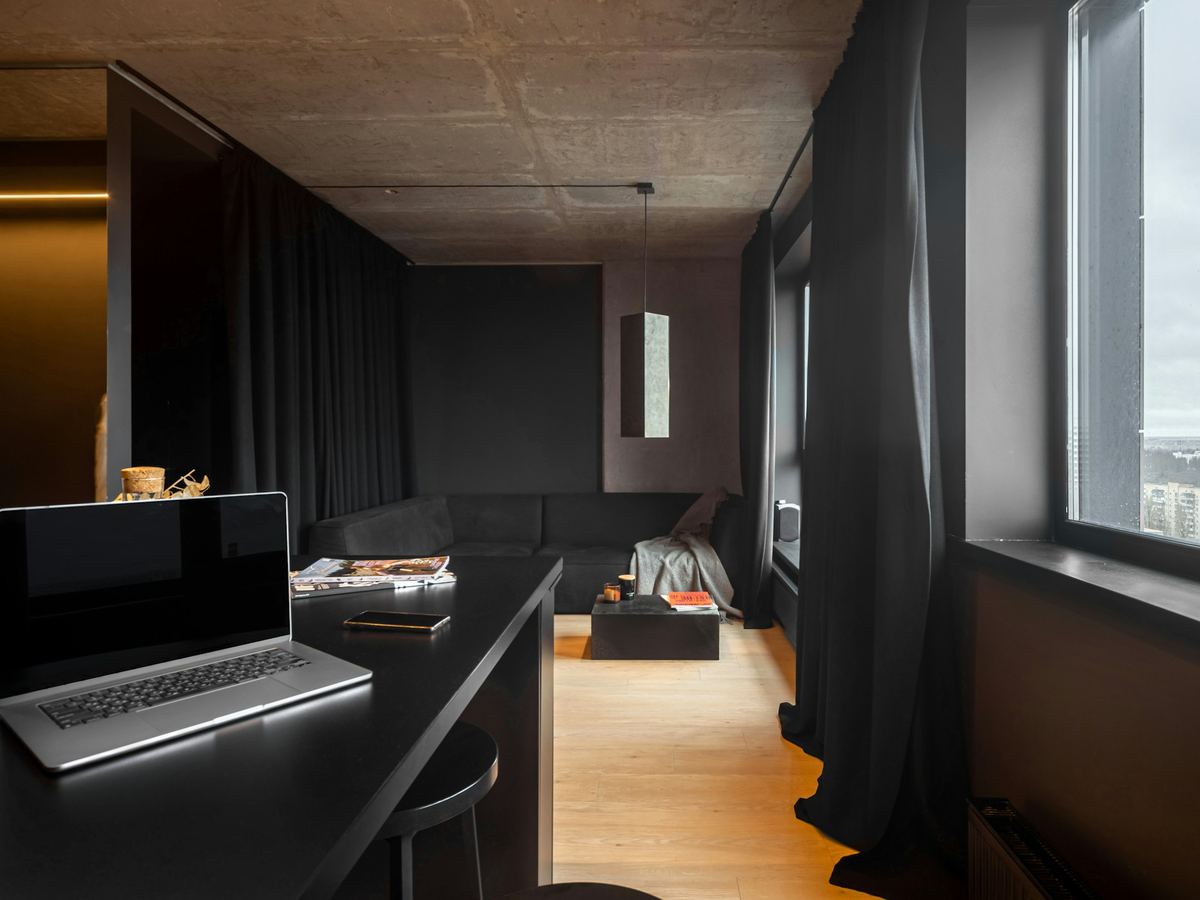 A dark, minimal apartment interior with a desk by the window