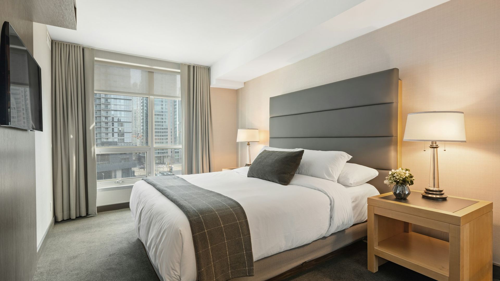 A made hotel bed with white linen beside a window onto the city